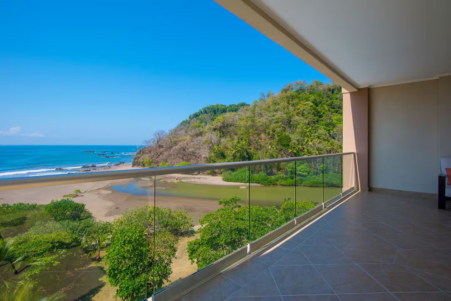 Tiled balcony with a glass railing overlooking a tropical beach, lagoon and forested hill