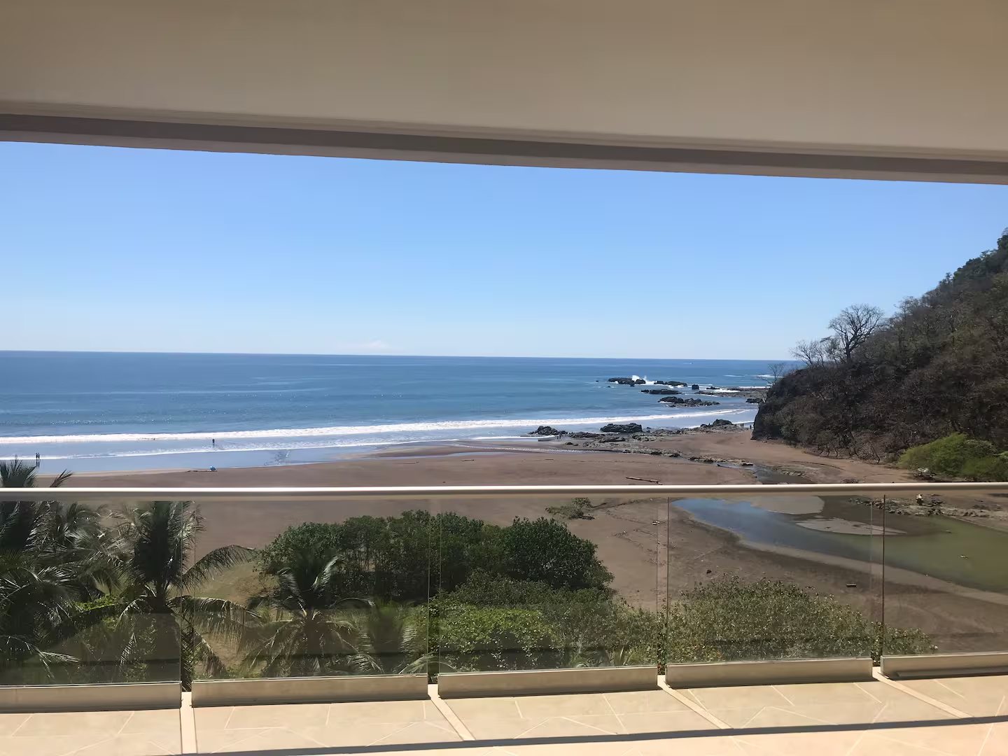 Balcony with glass railings overlooking a sandy beach, blue ocean and rocky cliffs