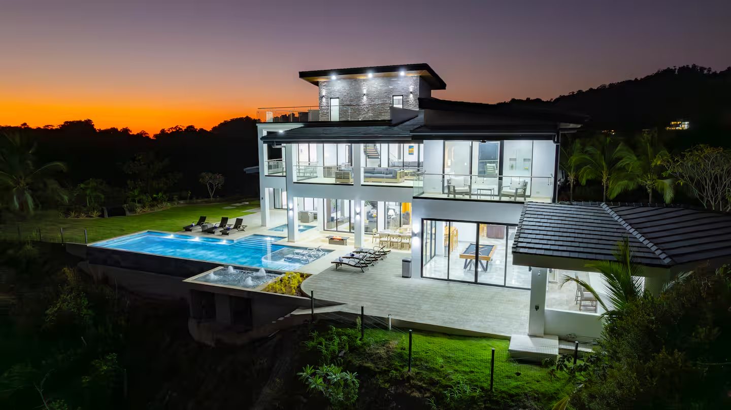 Modern multi-level villa with a lit pool and expansive glass walls at sunset