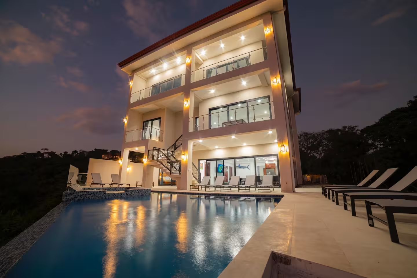 Modern three-storey villa with glass balconies and a lit infinity pool at dusk